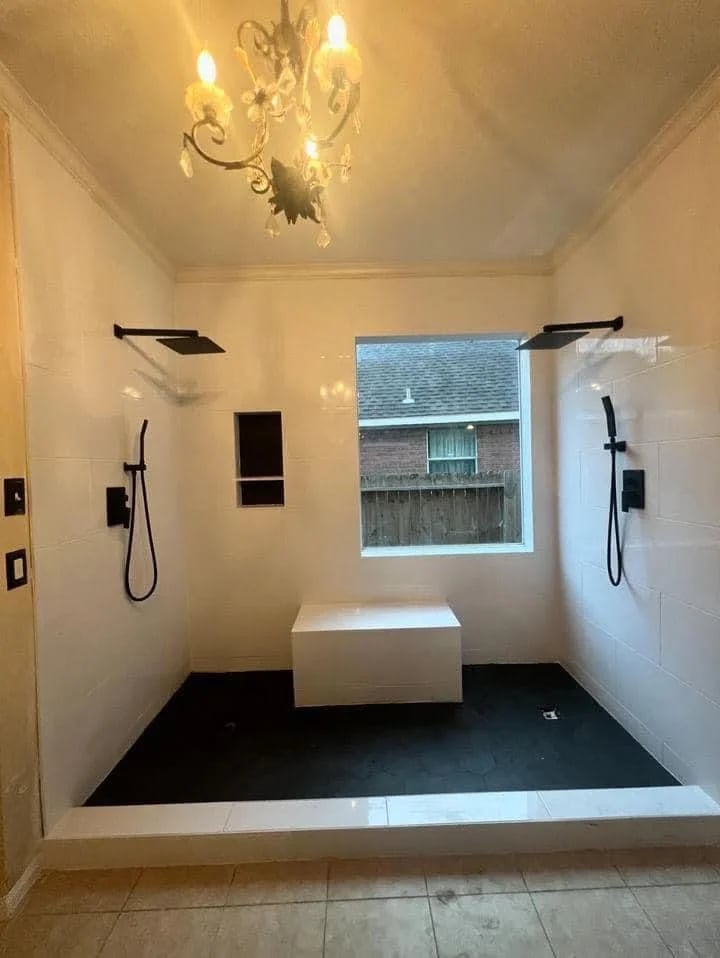 Open shower with two matte black rain showers and hand showers, white tile walls, a window, a tiled bench and a black hexagon tile floor.