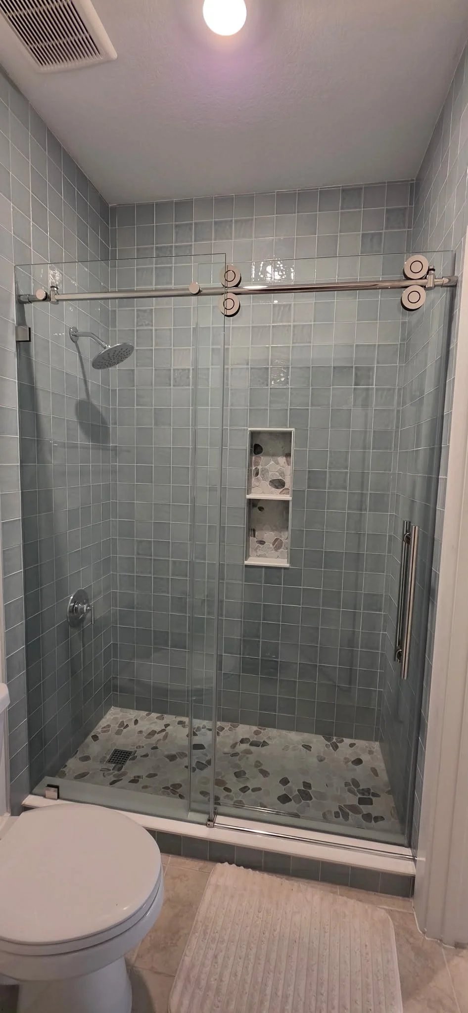Shower with glossy blue-grey square tile, a frameless sliding glass door, a three-shelf niche and a mixed pebble floor.