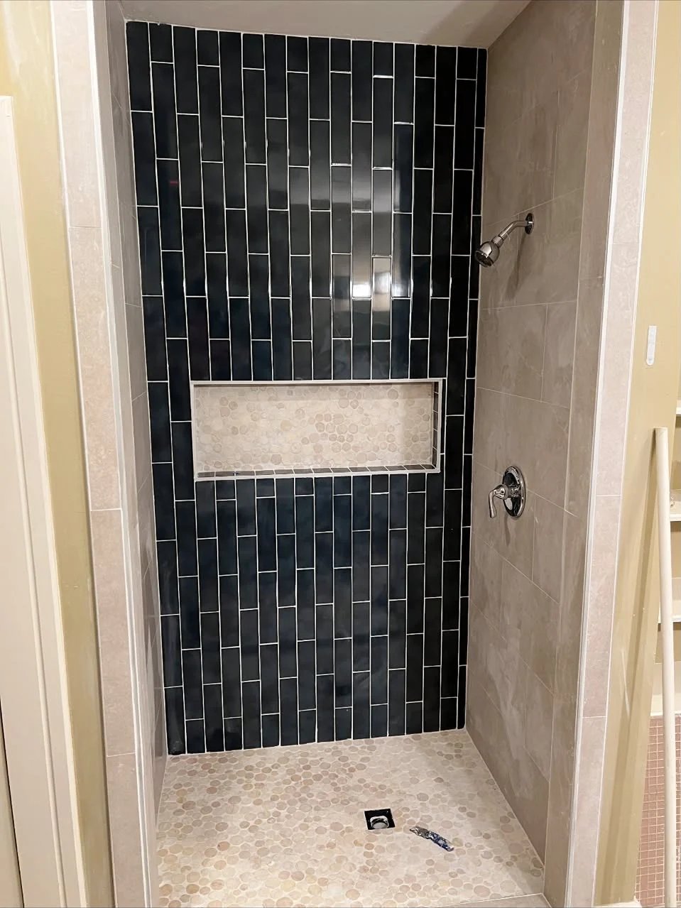 Shower with a navy blue vertical tile back wall, a beige mosaic niche, beige side walls, a chrome valve and a pebble floor.