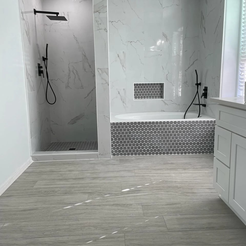 Bathroom with a walk-in shower and a separate tub in white marble-look tile, grey penny tile on the tub front, niche and shower floor, matte black fixtures and a grey wood-look floor.