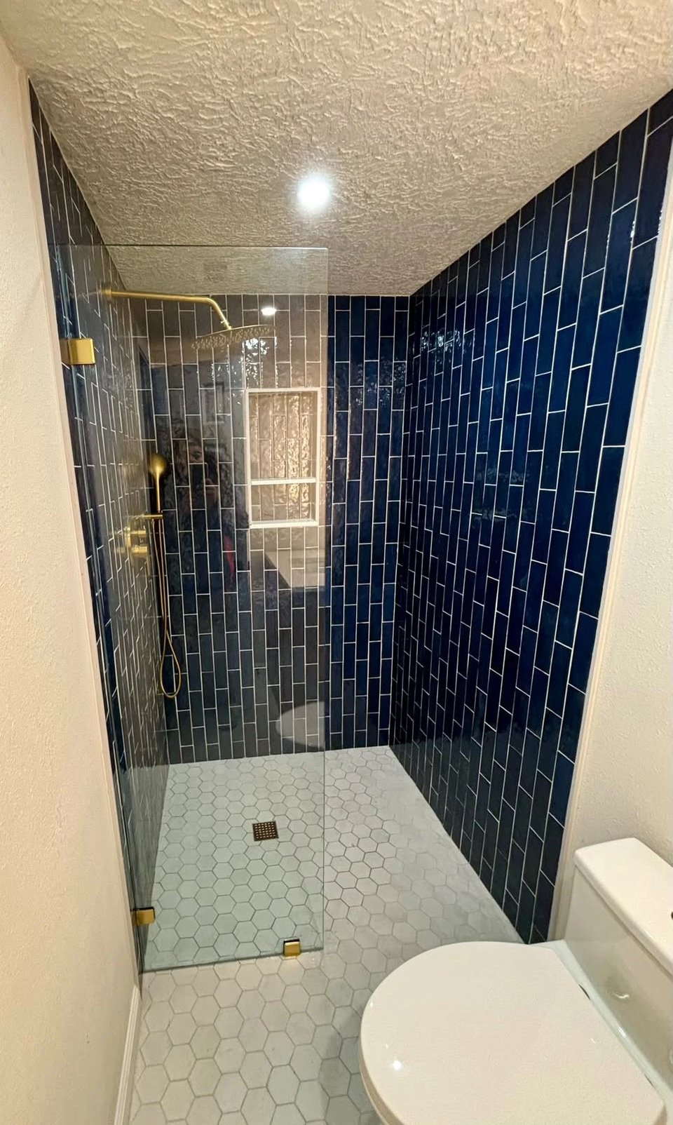 Walk-in shower with navy blue vertical subway tile, a brushed brass rain shower and hand shower, a frameless glass panel and a white hexagon tile floor.