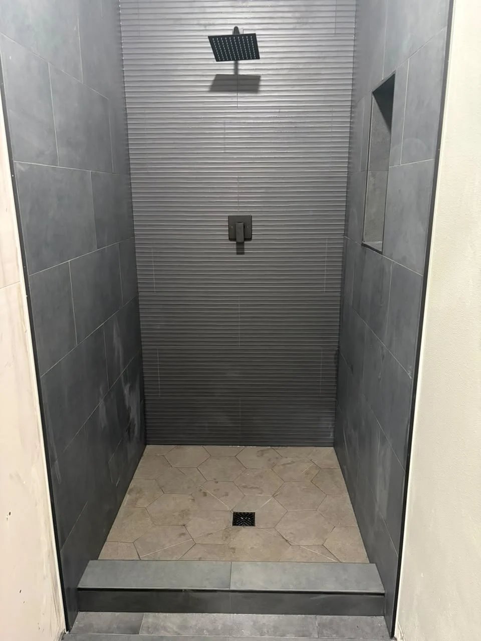 Shower with charcoal large-format tile, a ribbed accent wall, a matte black rain shower and valve, a niche and a large hexagon tile floor.
