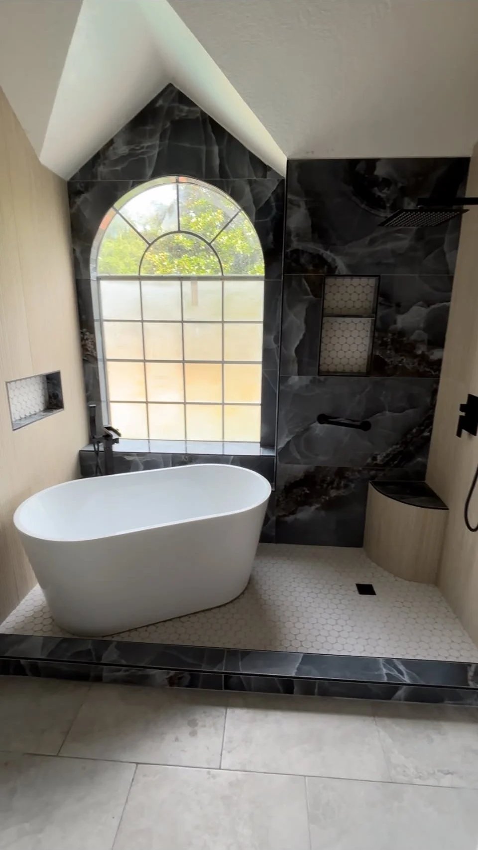 Bathroom with a white freestanding tub on a raised platform beneath an arched window, black marble-look tile walls, matte black fixtures and a white hexagon tile floor.