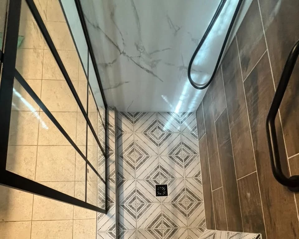 Shower with a black-and-white geometric patterned floor, wood-look plank tile, a white marble-look wall, a black-framed glass panel and a black grab bar.