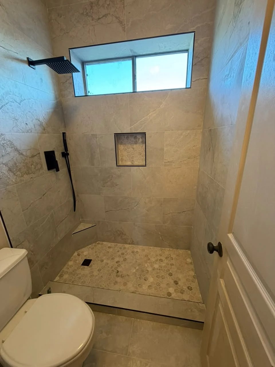 Shower with beige stone-look tile, a window above, a matte black rain shower and hand shower, a hexagon mosaic niche and floor, and a corner shelf.