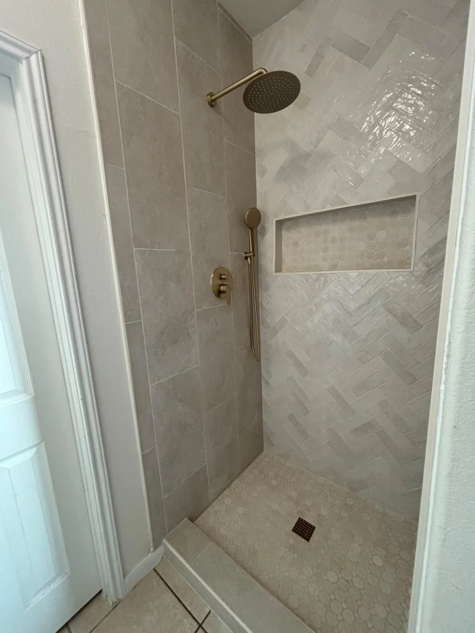 Shower with a glossy white herringbone wall, grey large-format tile, a brushed brass rain shower and hand shower, a long niche and a hexagon mosaic floor.