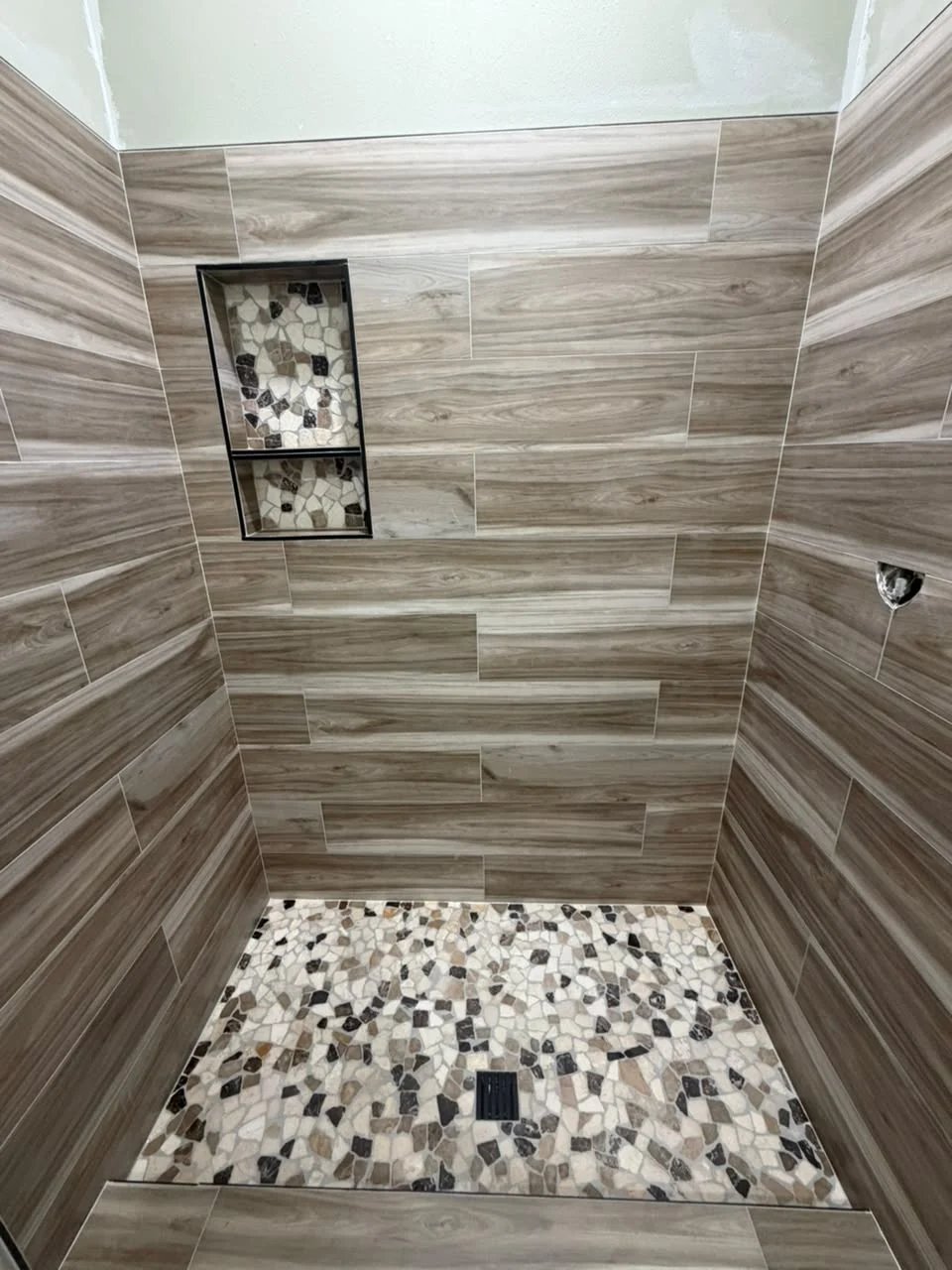 Shower with wood-look plank tile on the walls, a two-shelf niche lined in pebble mosaic and a floor in multicolored flat pebble mosaic.