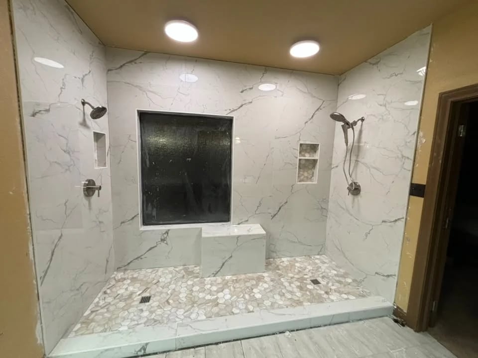 Large walk-in shower in white marble-look tile with two shower heads, a built-in bench, two niches and a river-rock pebble floor.