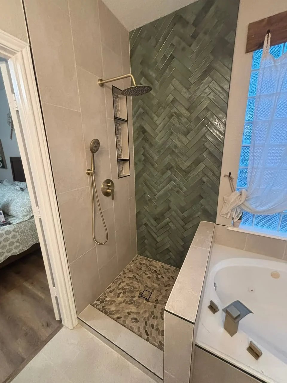 Shower with a sage-green herringbone accent wall, beige large-format tile, a brushed brass rain shower and hand shower, a tiled niche and a pebble floor, beside a jetted tub.
