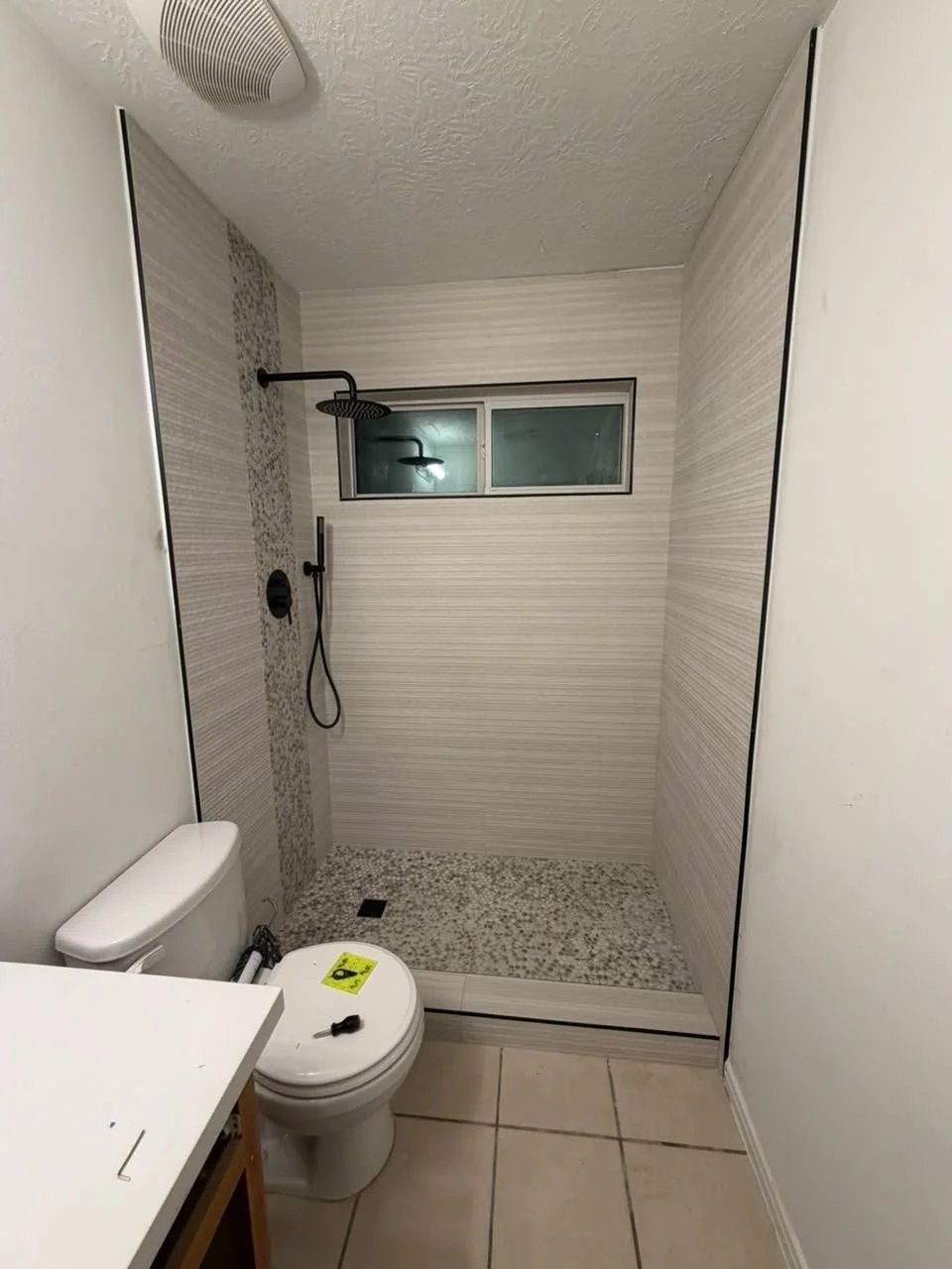 Shower with cream textured tile, a vertical pebble accent stripe, a window, a matte black rain shower and hand shower and a pebble floor.