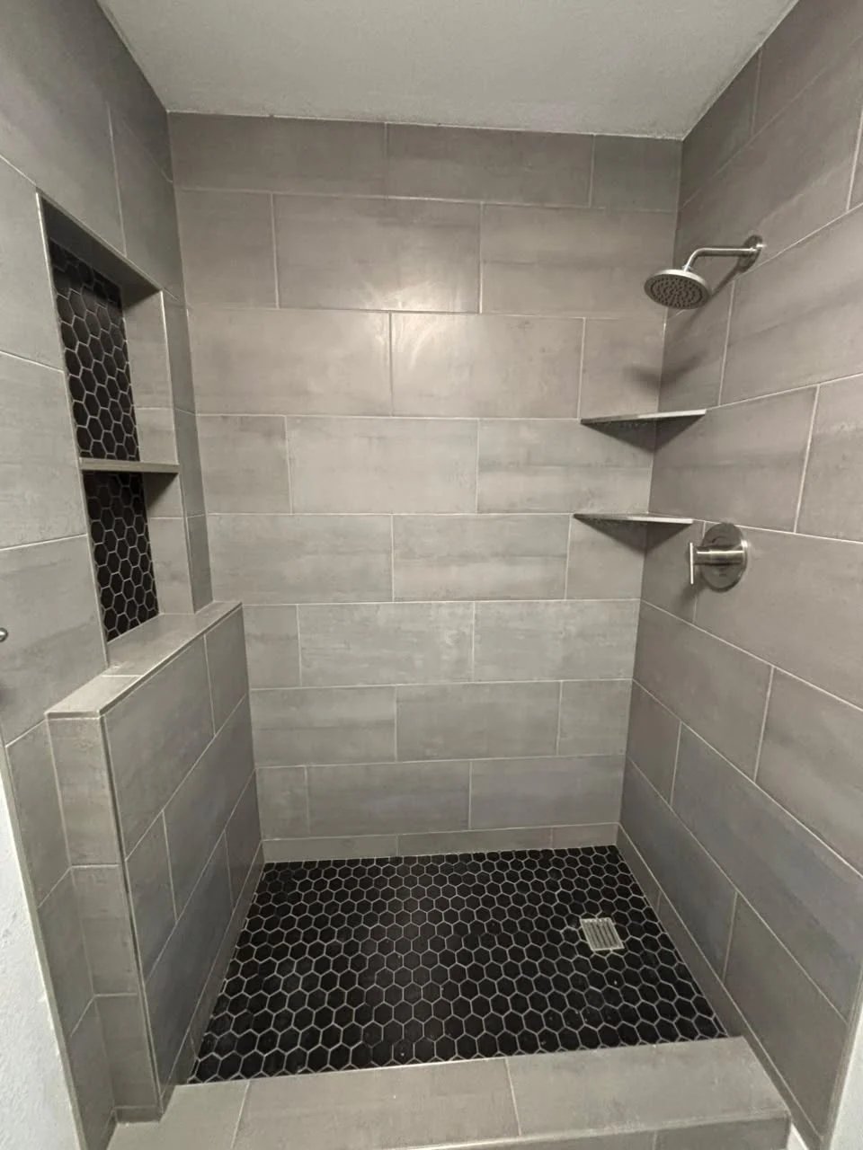 Shower with grey plank tile, a half wall, a two-shelf niche lined in black hexagon mosaic, corner shelves, brushed nickel fixtures and a black hexagon mosaic floor.